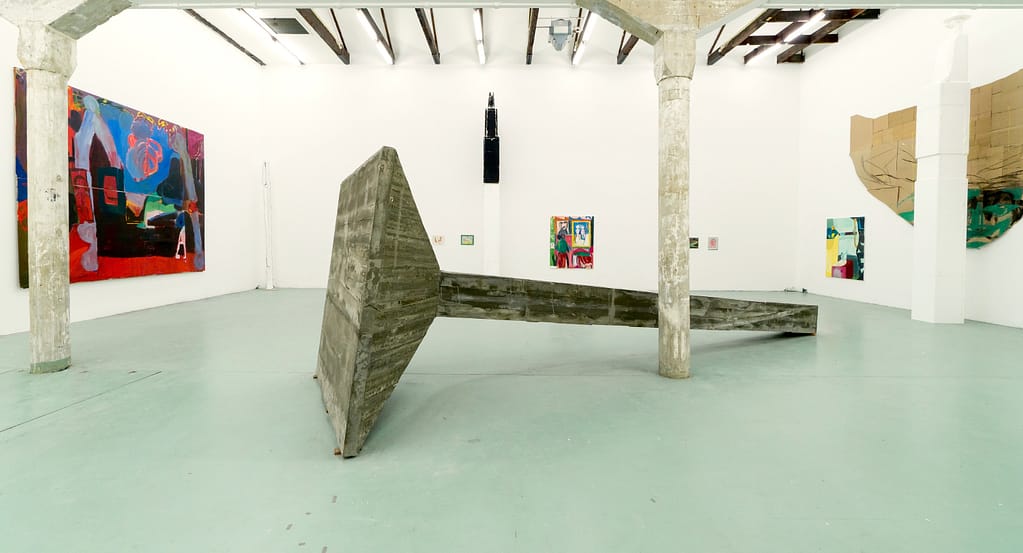 Contemporary art gallery installation in W139, featuring large abstract sculptures by Landzbergas and vibrant paintings by Axel Linderholm on white walls. A large, leaning concrete structure dominates the floor space, surrounded by colorful paintings with bold shapes.