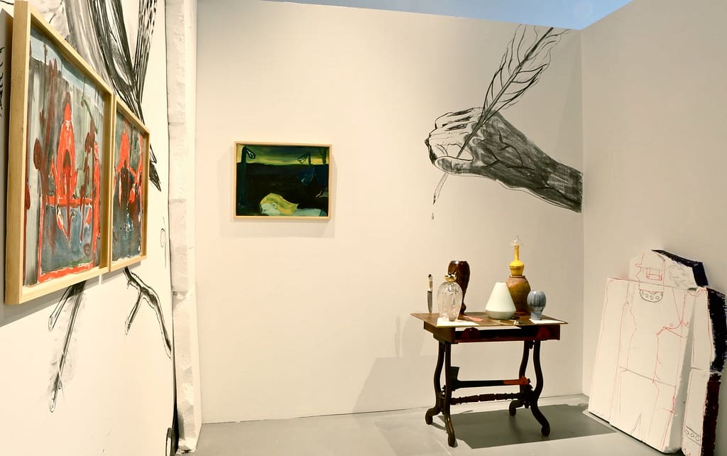 Art installation with dark atmospheric paintings, a drawing of a hand drawing, a sculpture of a table with pottery, and a styrofoam sculpture of a broken man in the Centre for Contemporary Art Vilnius.