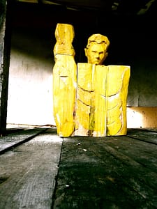 Yellow painted abstract wood sculpture resembling a human figure, with rough geometric features, standing in a rustic, dimly lit room with wooden floorboards. The figure's head is tilted, and its upper body is divided into block-like sections.