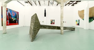 Contemporary art gallery installation in W139, featuring large abstract sculptures by Landzbergas and vibrant paintings by Axel Linderholm on white walls. A large, leaning concrete structure dominates the floor space, surrounded by colorful paintings with bold shapes.