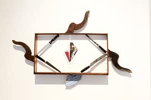 Contemporary art installation, featuring a wooden rectangular structure on a white wall. Four knives make a square within this rectangle, framing a cut up photo of a man.