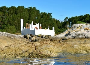Contemporary installation on a rock in Norway. The photo, taken from the water, shows a white castle with abstract towers and sculptures on the walls, both painted and of wood.