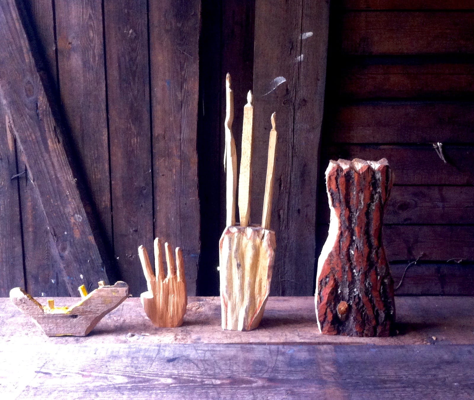 Abstract wood sculptures resembling hands, jaws and fists, with rough geometric features, standing in a rustic, dimly lit room with wooden walls.