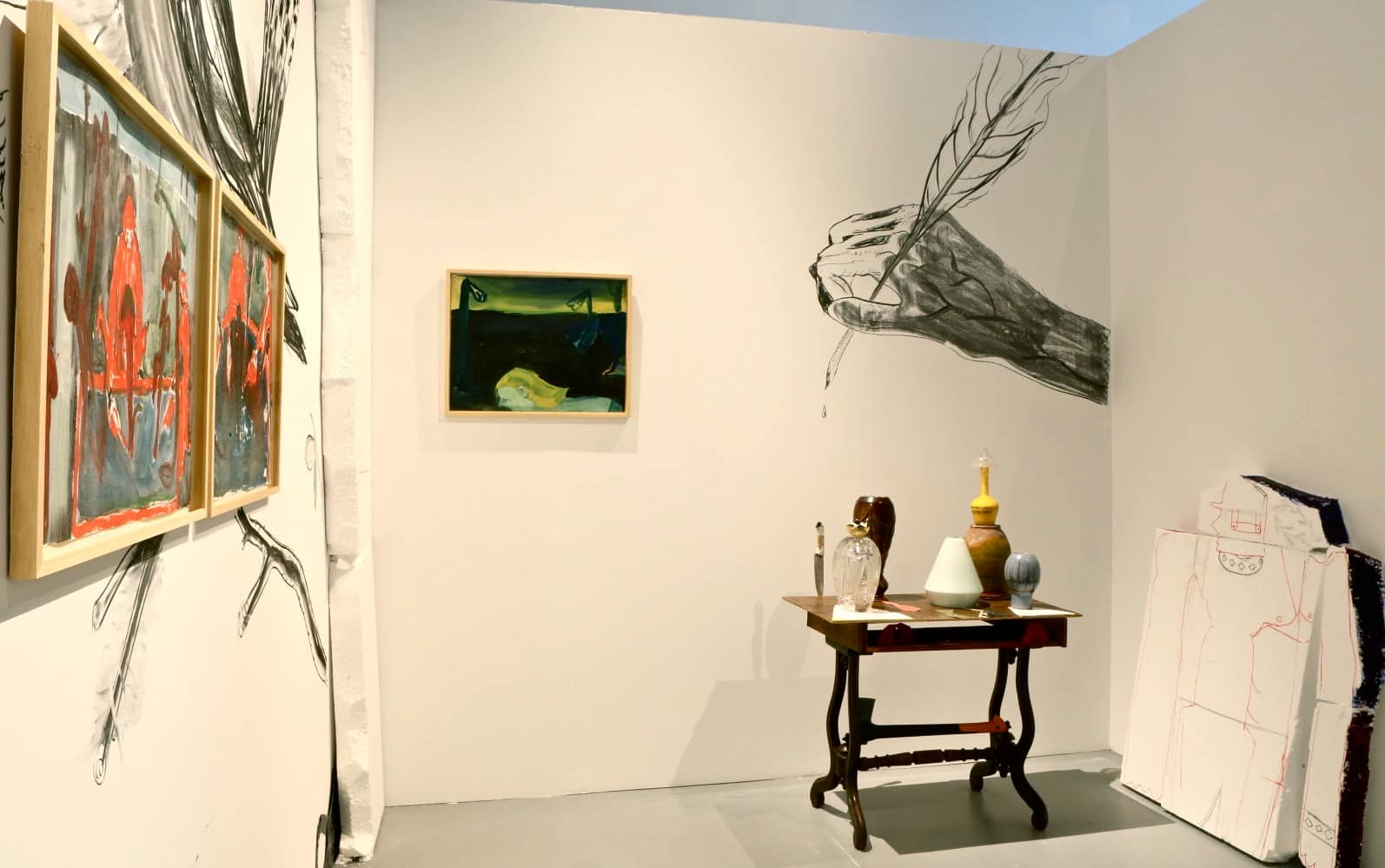 Art installation with dark atmospheric paintings, a drawing of a hand drawing, a sculpture of a table with pottery, and a styrofoam sculpture of a broken man in the Centre for Contemporary Art Vilnius.