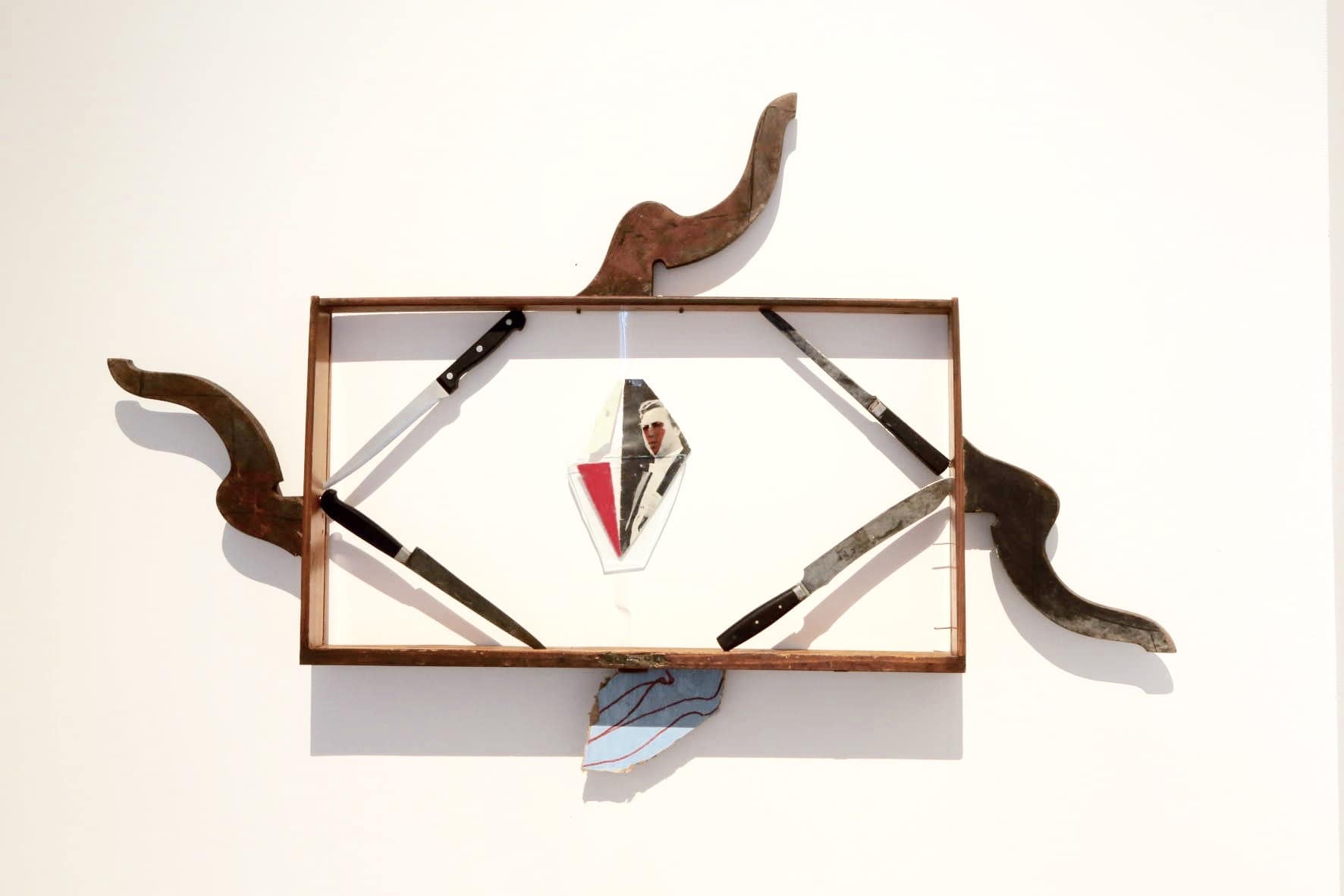 Contemporary art installation, featuring a wooden rectangular structure on a white wall. Four knives make a square within this rectangle, framing a cut up photo of a man.